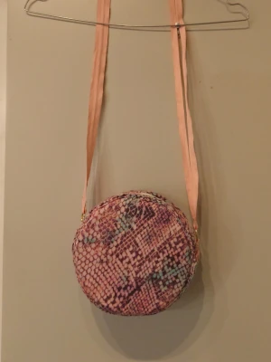 Rund axelväska med ormmönster - Säljer en rund axelväska med ormmönster i rosa, lila och turkosa toner. Väskan har ett mjukt tygmaterial och en ljusrosa axelrem. Stängs med dragkedja och har guldfärgade ringar vid remfästet. Perfekt för dig som gillar unika accessoarer. Den är sydd av mig av en återbrukad tjocktröja 