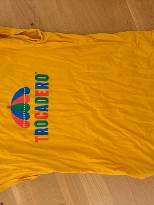 Gul Trocadero t-shirt med tryck - Säljer en gul t-shirt från Trocadero med stort färgglatt tryck på bröstet. T-shirten har klassisk rund hals och korta ärmar. Materialet är mjuk bomull, perfekt för chill dagar och sommarhäng. Färgen är riktigt poppig och sticker ut i mängden.