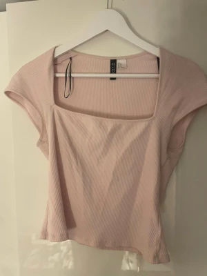 Ljusrosa ribbad topp från Divided - Säljer en ljusrosa ribbad topp från Divided med fyrkantig halsringning och korta ärmar. Toppen har en slim passform och är tillverkad i mjuk bomullsmix som känns skön mot huden. Perfekt att styla med jeans eller kjol för en fräsch look.