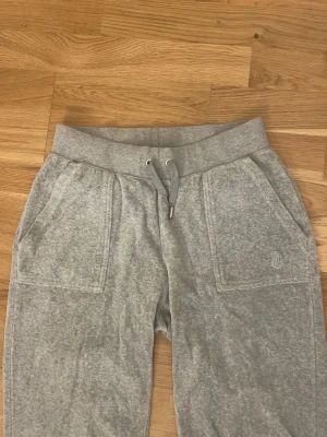Grå mjukisbyxor Juicy Couture XS - Grå mjukisbyxor från Juicy Couture i storlek XS. Byxorna har snörning i midjan, stora fickor fram och bak med knappdetaljer, samt ett broderat Juicy Couture-emblem på ena fickan. Mjukt material som är skönt att ha på sig.
