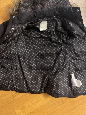 Svart pufferjacka med fuskpäls från Name It - Svart pufferjacka från Name It med mysig huva och fuskpäls. Insidan har ett prickigt mönster och jackan stängs med både dragkedja och knappar. Perfekt för kalla dagar och har praktiska fickor framtill.