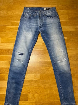 Slim fit blå jeans från Chasin' - Säljer ett par slim fit jeans från Chasin' i tvättad blå färg med slitningar och hål på benen. Jeansen har klassiska fem fickor, knappgylf och coola bakfickor med unik söm. Perfekt för dig som gillar en modern och avslappnad stil.