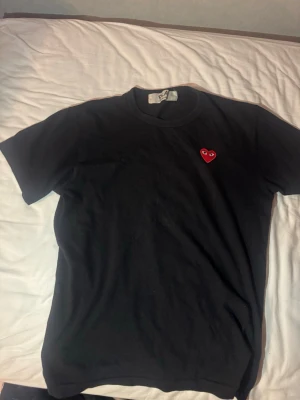  Comme des Garçons Play t-shirt (M) - Svart t-shirt från Comme des Garçons Play med klassisk rund hals och korta ärmar. På bröstet sitter det ikoniska röda hjärtat med ögon, broderat. T-shirten är gjord i mjuk bomull och har en clean, stilren vibe som är lätt att matcha.