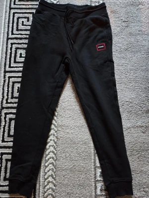 Svarta mjukisbyxor från Hugo - Svarta mjukisbyxor från Hugo med en röd logotyp-patch på vänster ben. Byxorna har snörning i midjan och ribbade muddar vid bensluten. Perfekta för chill eller häng med kompisar. Mjukt material som känns skönt mot huden.