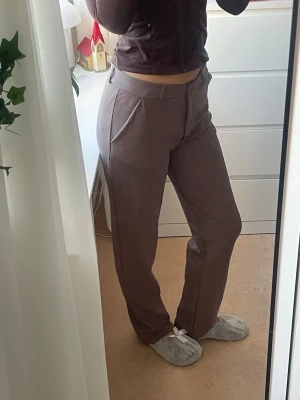 Bruna kostymbyxor med rak passform - Snygga bruna kostymbyxor från lager 157 i rak modell med midwaist. Byxorna har klassiska fickor, bälteshällor och stängs med knapp och dragkedja. Perfekta till en chill look med sneakers eller boots. Materialet är inte klassiskt kostym material, mer luftigt och stretchigt.  Lite store i storleken 