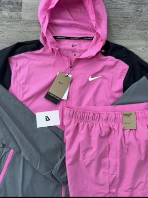 Nike truck suit - Bilden visar en rosa och grå Nike träningsjacka med huva och matchande rosa träningsbyxor, men inga skor finns med.