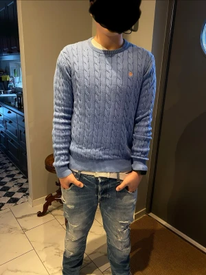 Ljusblå kabelstickad tröja Morris - Säljer en ljusblå kabelstickad tröja från Morris med rund hals och broderad logga på bröstet. Tröjan har lång ärm och ribbade muddar vid ärmslut och nederkant. Perfekt för lager-på-lager och snygg till jeans.