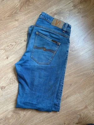 Blå jeans från Nudie Jeans 33/34 - Säljer ett par klassiska blå jeans från Nudie Jeans i storlek 33/34. Jeansen har fem fickor, snygga kontrastsömmar och Nudie-logga på bakfickan. Modellen har rak passform och är tillverkad i slitstarkt bomullsmaterial. Perfekt för dig som gillar tidlös denimstil.