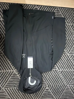 Svart vindjacka från Haglöfs XL - Svart vindjacka från Haglöfs i storlek XL, designad för kvinnor. Jackan har minimalistisk look, hel dragkedja framtill och justerbar huva. Perfekt för dig som vill ha en stilren och funktionell jacka till utelivet. Materialet är slitstark syntet.