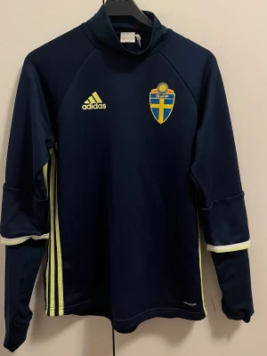 Mörkblå adidas Sverige träningströja - Säljer en mörkblå träningströja från adidas med Sveriges landslagsmärke på bröstet och gula detaljer. Tröjan har lång ärm, hög krage och gula ränder längs sidorna. Perfekt för dig som gillar fotboll och vill representera Sverige.