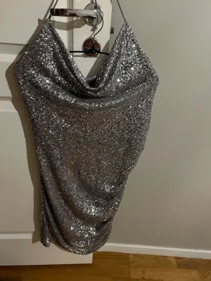 Silver paljettklänning från BikBok - Säljer en glittrig silverfärgad paljettklänning från BikBok med halterneck och draperad detalj på sidan. Klänningen är kort och har en snygg omlottkänsla framtill. Perfekt för dig som vill sticka ut! Använd en gång. 