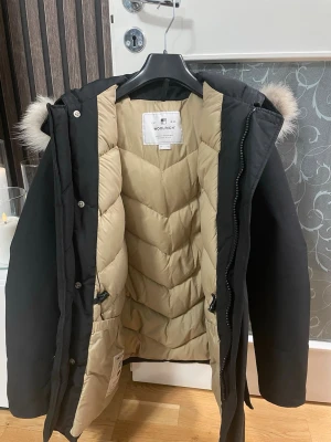 Svart parkas från Woolrich med päls - Säljer en svart parkas från Woolrich med beige quiltat foder och fluffig pälsdetalj på huvan. Jackan har dragkedja och knappar framtill, stora fickor och lång ärm. Perfekt för kalla dagar och riktigt snygg till vinterlooken. Skicka gärna frågor och bud!