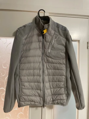 Grå pufferjacka från Parajumpers - Snygg grå pufferjacka från Parajumpers med quiltad framsida och rygg, samt släta ärmar i stretchigt material. Jackan har dragkedja framtill, bröstficka med dragkedja och den klassiska gula detaljen vid kragen. Perfekt för dig som vill ha en stilren och sportig look. Skicka gärna frågor och bud!