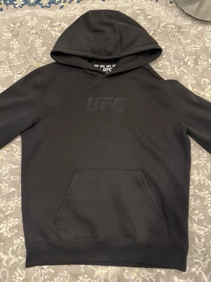 UFC Hoodie – Svart huvtröja med stort ryggtryck - Snygg och bekväm UFC-huvtröja i svart. Diskret UFC-logga på bröstet och ett stort, detaljerat tryck på ryggen med “Ultimate Fighting Championship – Las Vegas, Nevada”. Tröjan har huva och magficka, perfekt för både vardag och träning. Använd och i gott skick.