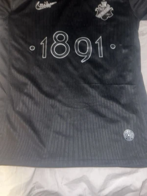 Svart AIK fotbollströja 1891 Smokinglirare edition  - Säljer en svart AIK fotbollströja med diskret randigt mönster och trycket '1891' på bröstet. Tröjan har klubbmärket och sponsorlogga framtill, samt en rund hals. Perfekt för dig som vill visa ditt stöd för AIK. Smokinglirare edition 
