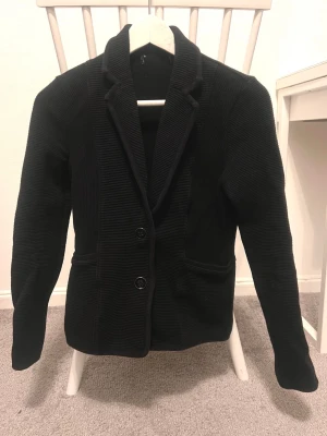 Svart manchesterkavaj med knappar - Säljer en svart kavaj i manchestertyg med två knappar framtill och klassisk krage. Jackan har lång ärm och ribbad struktur som ger en snygg och stilren look. Perfekt för dig som vill ha en dressad men ändå chill stil.