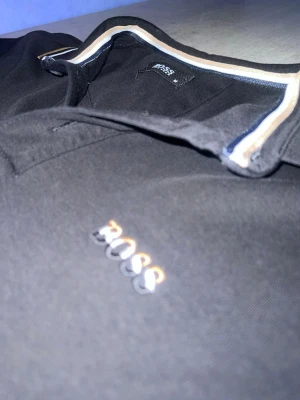 Svart piké från BOSS med gulddetaljer - Svart pikétröja från BOSS med broderad logga på bröstet. Kragen och ärmsluten har snygga ränder i vitt, guld och marinblått. Klassisk pikémodell med korta ärmar och knappslå framtill. Perfekt för en clean och stilren look.