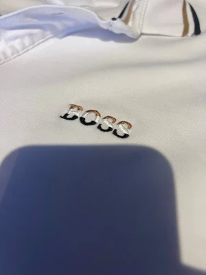 Vit polotopp från BOSS - Snygg vit polotopp från BOSS med broderad logga på bröstet. Kragen och ärmsluten har stilrena ränder i svart och beige. Toppen har korta ärmar och klassisk krage med knappar. Perfekt för en clean och sportig look.