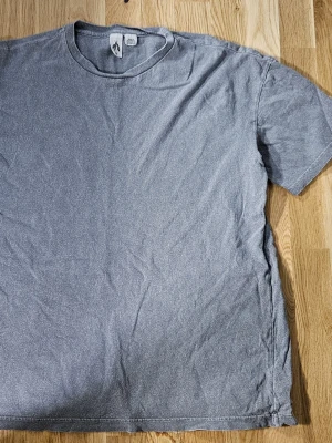 Grå t-shirt med rund hals M - En enkel grå t-shirt i storlek M med rundad hals och korta ärmar. Passformen är rätt pösig och skön. T-shirten är tillverkad i mjuk bomull och har en neutral look som funkar till det mesta.