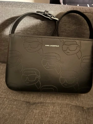 Svart axelväska från Karl Lagerfeld - Snygg svart axelväska från Karl Lagerfeld med präglade motiv av Karl Lagerfelds ansikte över hela väskan. Väskan är tillverkad i skinn och har en justerbar axelrem samt dragkedja upptill. Insidan är ljus och har fack för småsaker. Perfekt för dig som gillar stilrena designer kommer i dustbag 