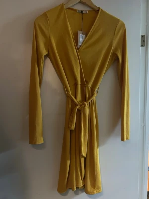 Gul omlottklänning från NLY Trend - Snygg gul omlottklänning från NLY Trend i ribbad polyester. Klänningen har lång ärm, v-ringad hals och ett knytband i midjan som ger en schysst siluett. Kort längd och mjukt, stretchigt material som sitter bekvämt. Perfekt för dig som gillar färg och stil.