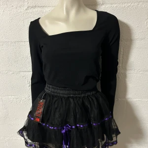 Svart volangkjol med lila detaljer Poizen - Kort svart kjol från Poizen med flera lager volang i mesh och lila banddetaljer. Kjolen har resår i midjan. Märke Poizen. Helt ny.