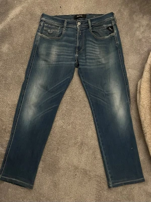 Replay blå jeans, midja 32 längd 30 - Säljer ett par snygga blå replay anbass hyperflex jeans med klassisk femficksdesign och diskret slitning framtill. Jeansen har raka ben och normal passform, med tydliga sömmar och Replay-logga på fickan. Perfekt för dig som gillar stilrena och bekväma jeans.