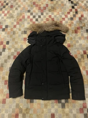 Svart dunjacka från Canada Goose - En knapp saknas där nere men en enkel fix. Orkar inte med Canada Goose.              Säljer en svart dunjacka från Canada Goose med pälskant på huvan och klassisk logga på ärmen. Jackan har lång ärm, knappar och dragkedja framtill. Perfekt för kalla vinterdagar och har en snygg, normal passform.