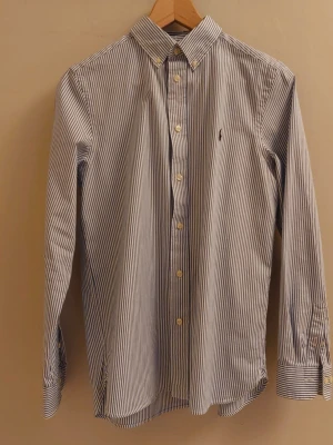 Oxford skjorta ungdom - Snygg randig skjorta från Polo Ralph Lauren i slim fit-modell. Skjortan är vit och blå med klassisk krage, knappar framtill och det ikoniska broderade logot på bröstet. Perfekt för dig som gillar stilrena och tidlösa plagg. Storlek 18, passar en ca 14 åring.