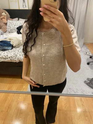 Beige kortärmad kofta med pärlknappar - Säljer en beige, kortärmad kofta med rund hals och pärlknappar framtill. Koftan är stickad och har en mjuk, lite fluffig känsla. Perfekt att slänga på över en topp för en chill och snygg look. Passar dig som gillar neutrala färger och enkel stil.