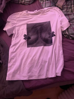 Vit t-shirt med tryck i storlek S - Säljer en vit t-shirt i storlek S med ett stort svart tryck på bröstet som föreställer en grafisk bild med tejpkryss. T-shirten har rund hals och korta ärmar. Materialet är polyester och elastan vilket gör den mjuk och lite stretchig.