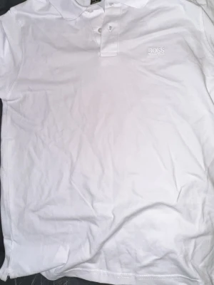 Vita t-shirts från BOSS - Två stilrena vita t-shirts från BOSS. Ena är en klassisk polo med krage och knappar, den andra har rund hals och tryckt BOSS-logga på bröstet. Båda är gjorda i mjuk bomull och har korta ärmar. Perfekt för dig som gillar enkel och clean stil.