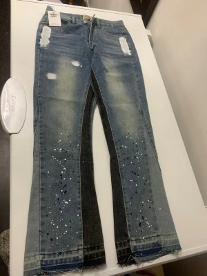 Gallery dept - Snygga grå jeans med vita och svarta färgstänk längs benen och slitna detaljer upptill. Byxorna har en rak passform och råa kanter vid bensluten. Materialet är klassisk denim och färgen är ljusgrå med inslag av svart längs sidorna.