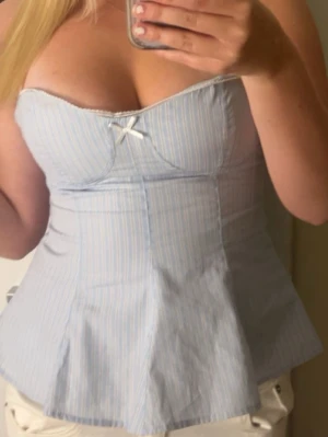 Blåvit randig korsett med rosett - Säljer en söt korsettopp i ljusblått och vitt med tunna ränder. Toppen har en liten vit rosett framtill och är axelbandslös med en figurnära passform. Perfekt för dig som gillar romantisk och feminin stil. 