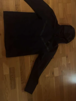 Nike tech  - Svart Nike tech med stilren design och huva. Hoodien har långa ärmar och dragkedja framtill, samt diskreta detaljer på bröstet. Perfekt för blåsiga dagar och enkel att matcha med olika outfits.
