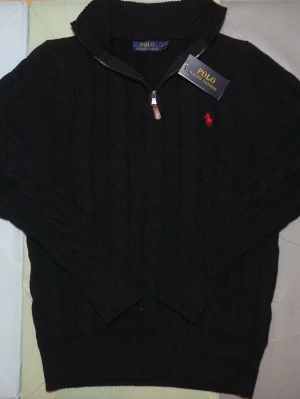 Svart stickad tröja Polo Ralph Lauren - Svart kabelstickad tröja från Polo Ralph Lauren med halv dragkedja och ribbad krage. Klassisk röd logga broderad på bröstet. Tröjan har långärmade och är gjord i mjukt stickat material, perfekt för kyliga dagar.