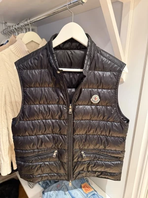 Moncler Gui väst - Snygg svart dunväst från Moncler med glansig finish och klassisk logotyp på bröstet. Västen har två dragkedjeförsedda fickor framtill och hög krage. Perfekt att slänga över en hoodie eller tröja för extra värme och stil. Västen är i övrigt mycket bra skick Den är i storlek s/2. Har tyvärr inte kvar kvittot så där av priset. 