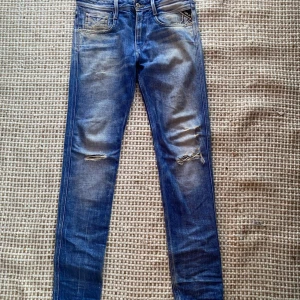 Replay Anbass blå slim jeans 29 - Säljer ett par Replay Anbass jeans i blå tvätt med slitningar och hål på benen. Slim fit med klassiska fem fickor och snygga detaljer på bakfickorna. Jeansen har dragkedjegylf och är tillverkade i stretchig bomullsdenim.