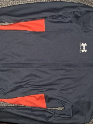 Under Armour Fleece - Snygg svart träningsjacka från Under Armour med dragkedja och röda detaljer på sidorna. Jackan har en sportig passform och är tillverkad i ett lätt och stretchigt material som passar perfekt till träning och aktivitet.