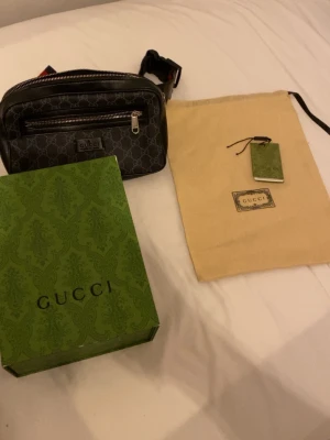 Svart axelväska  - Snygg svart axelväska med klassiskt GG-mönster och silverfärgade dragkedjor. Väskan har justerbar axelrem i rött och blått samt flera fack framtill. Kommer med originalbox, dustbag och tag.