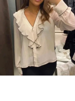 Vit blus med volang och v-ringning - Säljer en vit blus med djup v-ringning och stora volangdetaljer framtill. Blusen har långärmad design och är tillverkad i ett lätt, något transparent tyg. Perfekt för dig som gillar en romantisk och trendig stil.