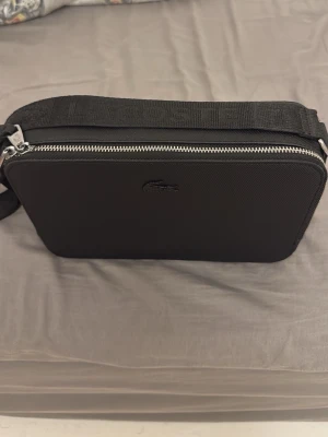 Lacoste väska - Säljer en stilren svart axelväska från Lacoste med justerbar rem där märket är broderat. Väskan har en silverfärgad dragkedja och ett minimalistiskt Lacoste-logo på framsidan. Insidan har ett extra fack med dragkedja. Perfekt för att bära dina essentials. 