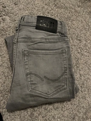 Grå skinny jeans från Jack and Jones  - Snygga grå jeans från Jack and Jones i en skinny modell. Jeansen har klassisk femficksdesign, svarta detaljer och ett svart Jack and Jones bak i midjan. Perfekta för dig som gillar en tajt passform och stilren look.