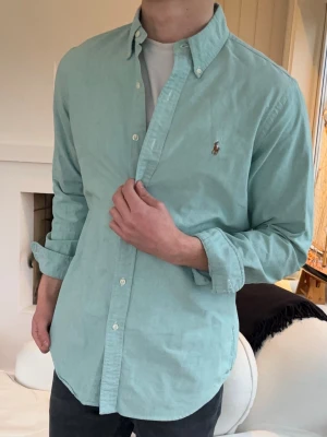 Mintgrön skjorta från Ralph Lauren - Stilren mintgrön skjorta från Ralph Lauren med klassisk button-down krage och broderad logga på bröstet. Skjortan har långa ärmar och vita knappar, perfekt för en avslappnad men snygg look.