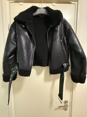 Svart skinnjacka med bälte och foder - Snygg svart skinnjacka med mjukt teddyfoder, bälte i midjan och dragkedjor på fickor och ärmar. Jackan har en croppad passform och detaljer i sidorna och vid nacken.
