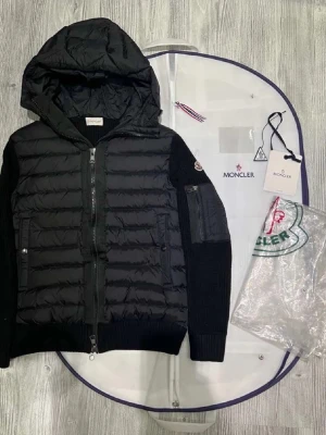 Svart dunjacka med huva från Moncler - Snygg svart dunjacka från Moncler med stickade ärmar och huva. Jackan har quiltad front, dragkedja och Moncler-logga på ärmen. Perfekt för kyliga dagar och ger en stilren look.