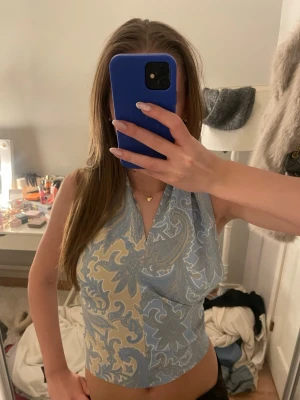 Blå och beige omlott halterneck topp - Snygg halterneck topp i blått och beige med paisleymönster. Toppen har omlottknytning i nacken och öppen rygg, vilket ger en trendig och somrig vibe. Perfekt att styla med jeans eller kjol för en cool look.