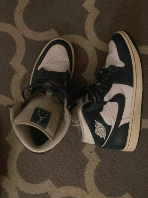 Nike Air Jordan 1 svartvita sneakers - Säljer ett par klassiska Nike Air Jordan 1 sneakers i svart och vitt. Skorna har hög modell med snörning, ikonisk swoosh på sidan och Air Jordan-logga vid ankeln. Ovandelen är i skinn och sulan är vit med svart yttersula. Perfekta för dig som gillar streetwear.