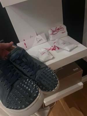 Blå mocka sneakers från Louboutin - Säljer ett par blå sneakers från Christian Louboutin i mocka med coola nitar över tån. Skorna har snörning och vit platt sula. Perfekta för dig som vill sticka ut med en edgy och lyxig stil. Snören box och extra nitar används 3 gånger så är väldigt fina säljer då dom ej passar längre 