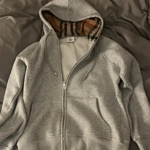Grå hoodie från Burberry med dragkedja - Säljer en grå hoodie från Burberry i storlek M. Tröjan har dragkedja, snörning i huvan och den klassiska Burberry-rutiga insidan av huvan. Tillverkad i mjuk bomull och har två fickor framtill. Perfekt för dig som gillar stilrena och exklusiva plagg.